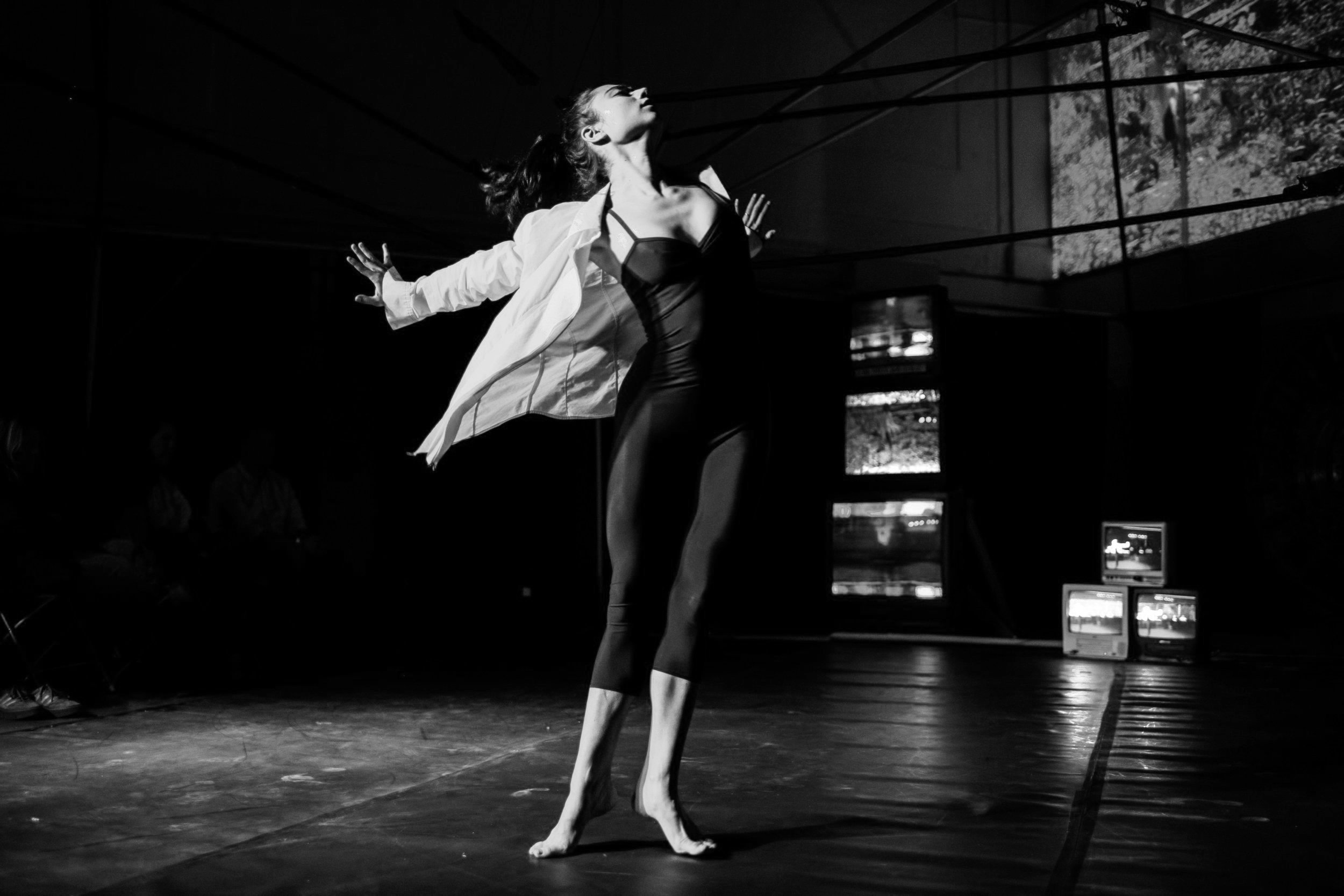A dancer performing on stage in a black leotard and white shirt with her arms outstretched and head tilted back, surrounded by stage lighting and monitors.