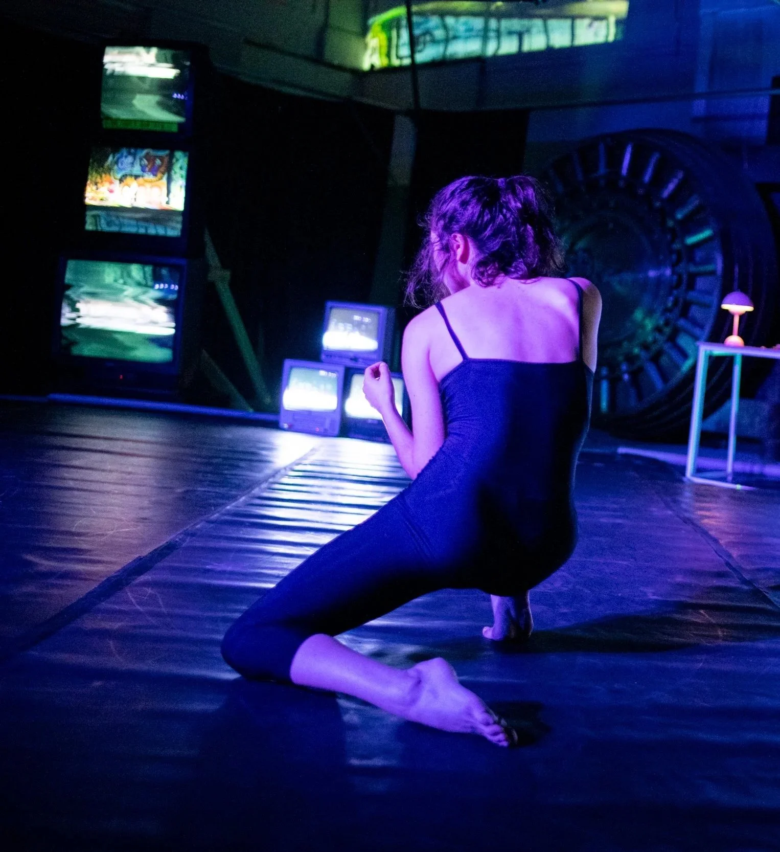 A woman practicing yoga on a black floor in a dimly lit room with colorful screens and a large decorative wall in the background.