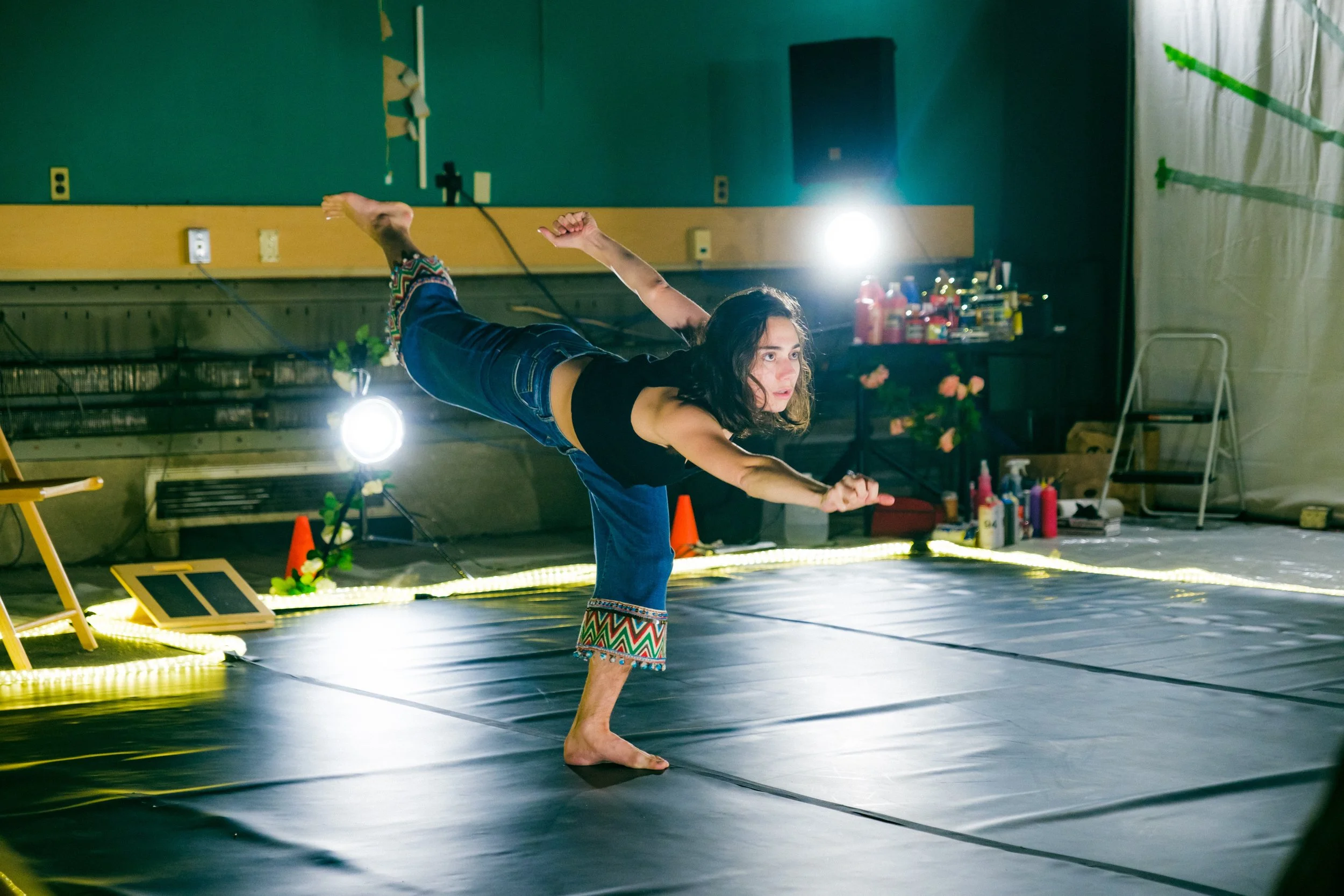 A woman practicing yoga or a balancing pose indoors on a mat, with bright lighting and colorful decor and supplies in the background.