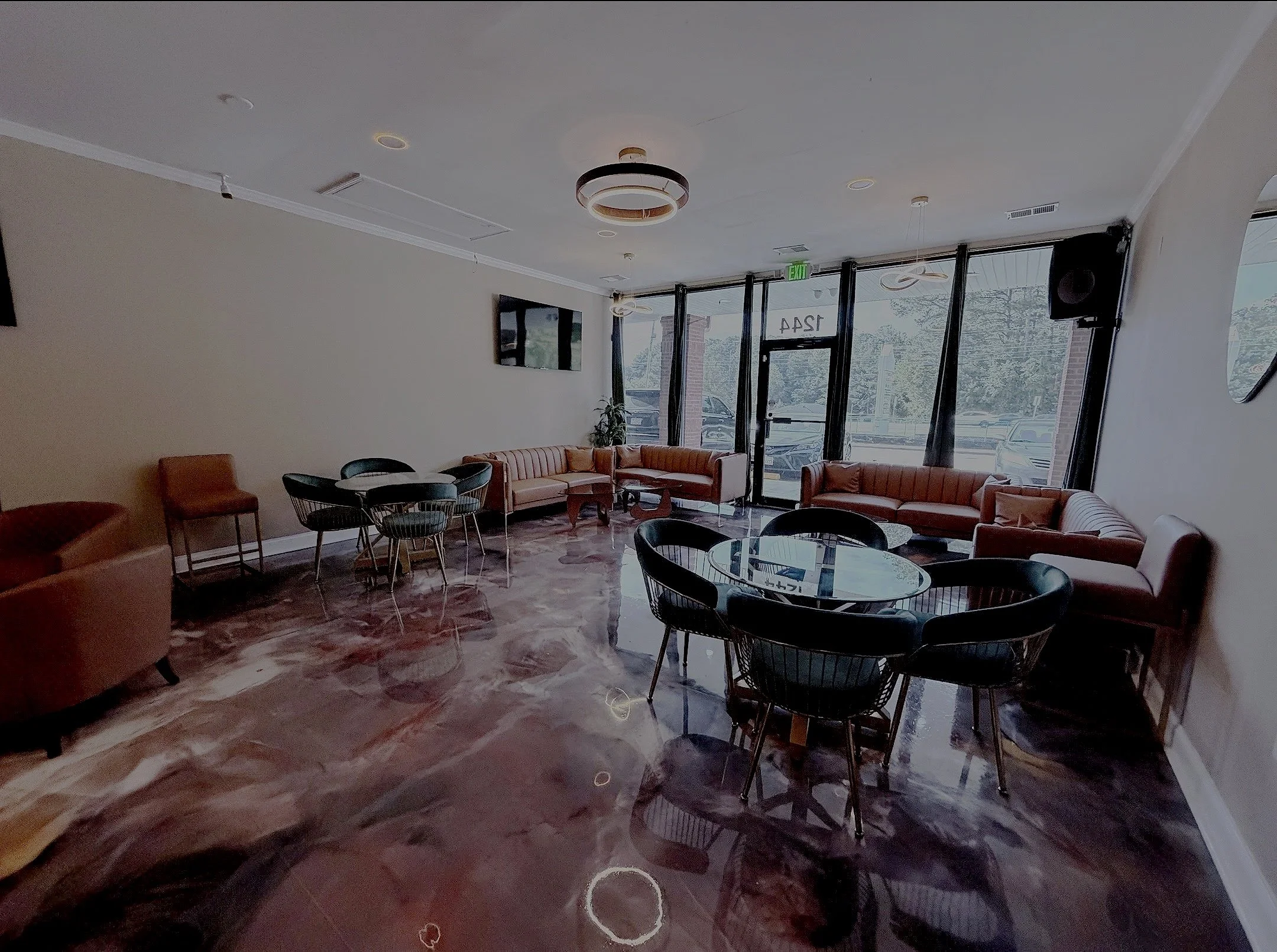 Interior of a modern cafe with pink couches, black and gold chairs, marble flooring, wall-mounted television, large glass windows with views of parking lot, and contemporary ceiling lights.