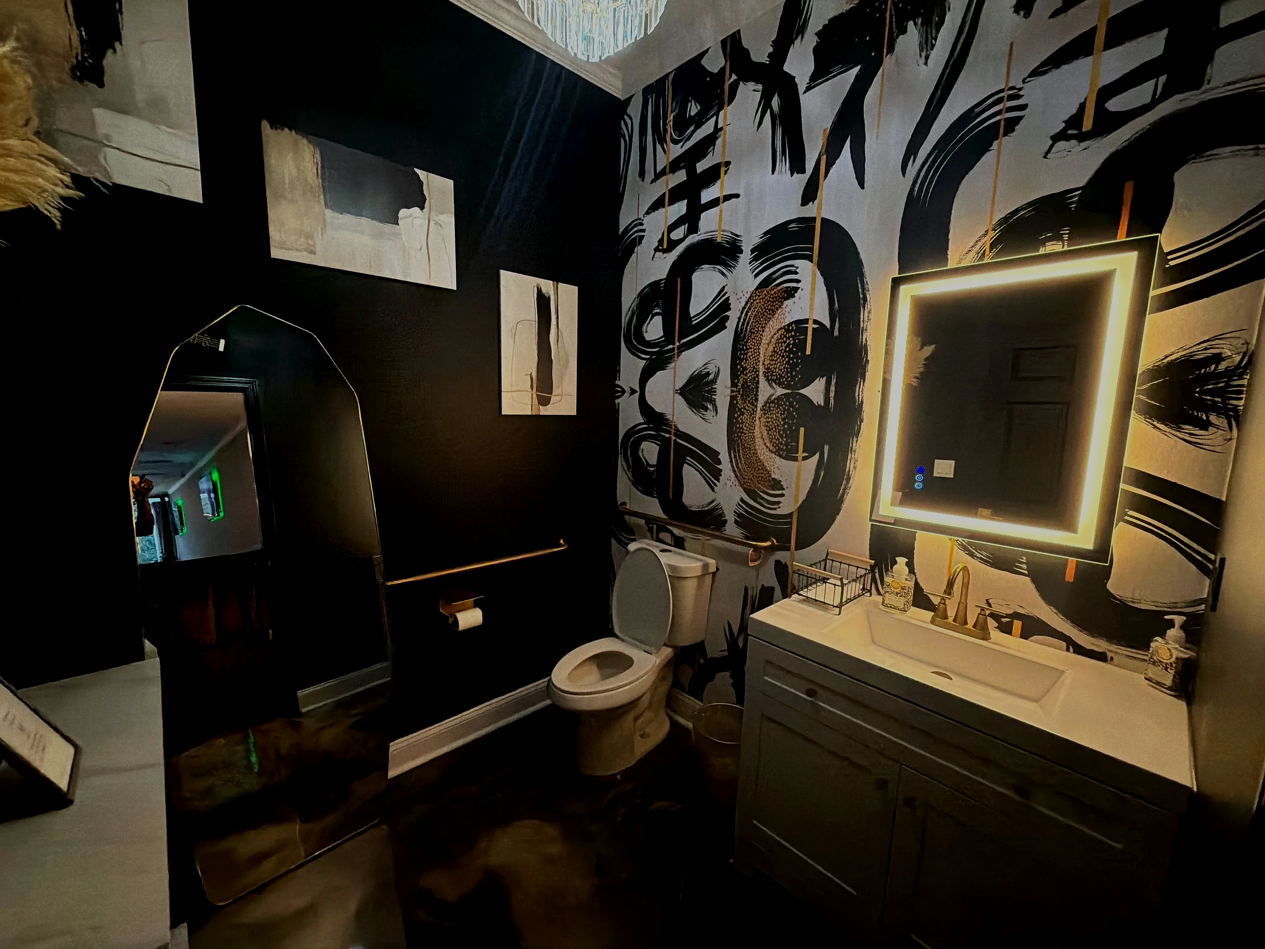 Bathroom with black and white abstract wall art, a mirror with LED lighting, a white vanity, a toilet, and various toiletries.