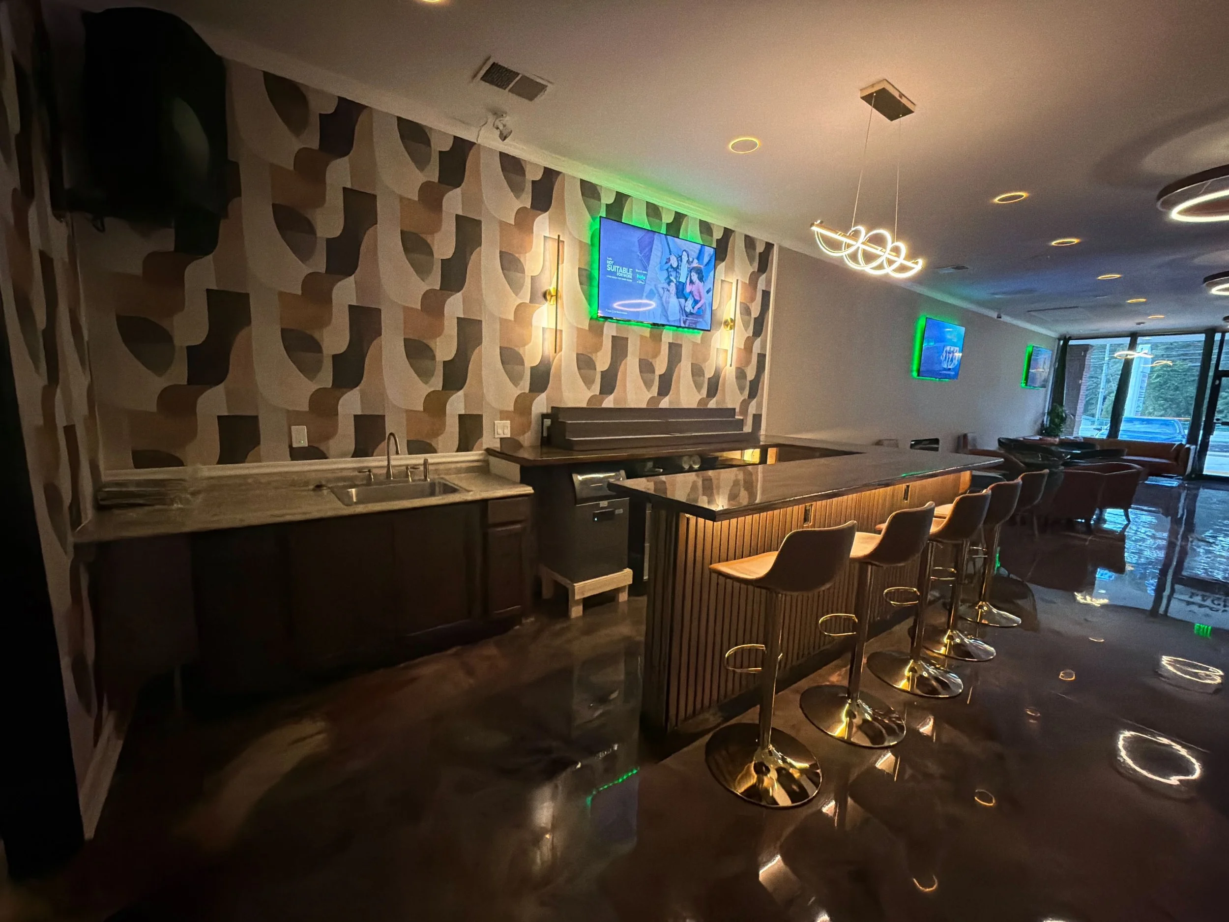 Interior of a modern bar with a patterned accent wall, multiple TV screens, illuminated hanging light fixtures, and a row of bar stools alongside a high-top counter.