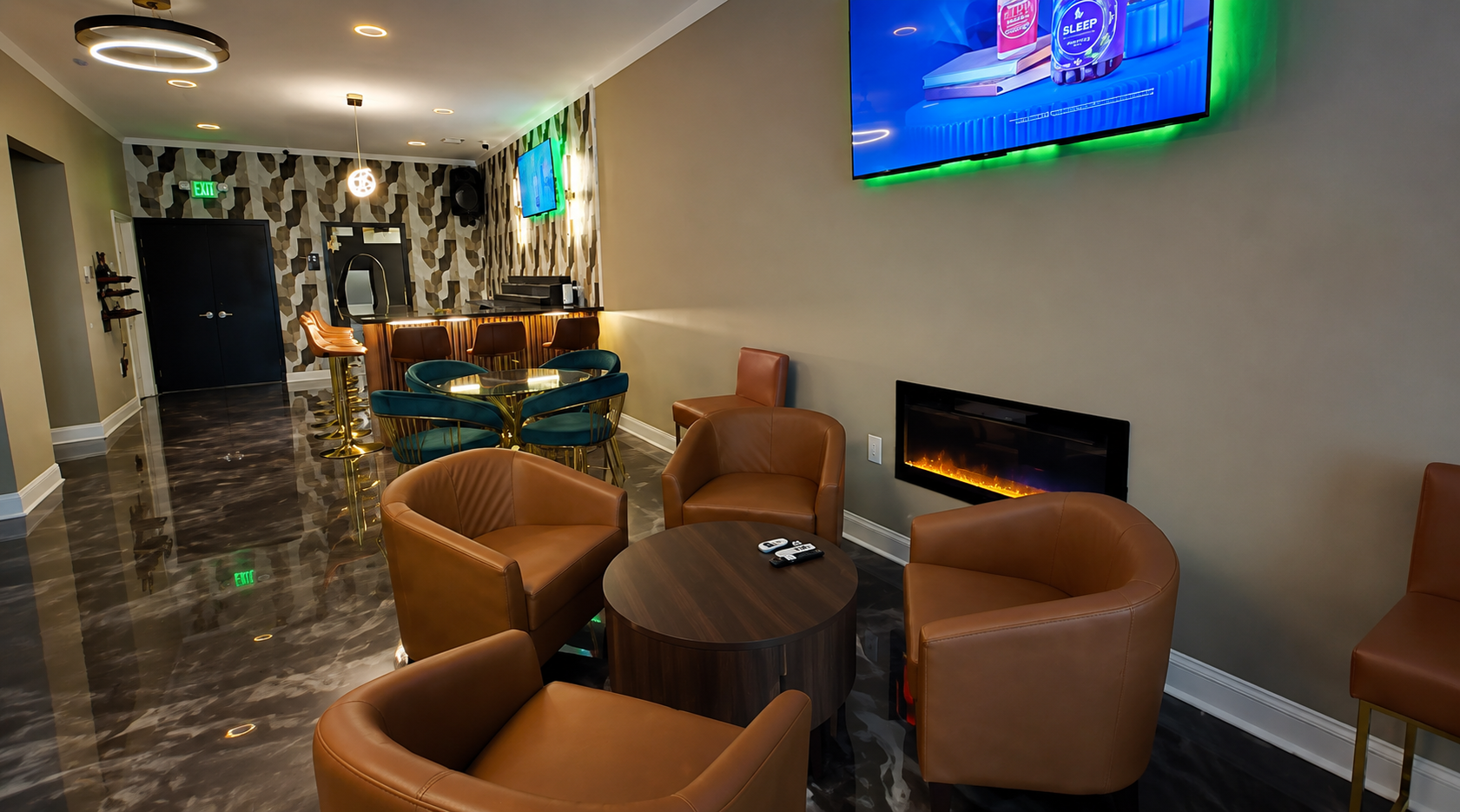 Living area with tan leather chairs arranged around a small wooden table, a wall-mounted electric fireplace, a flat-screen TV on the wall, a bar with barstools, a patterned accent wall, and a flat-screen TV overhead.