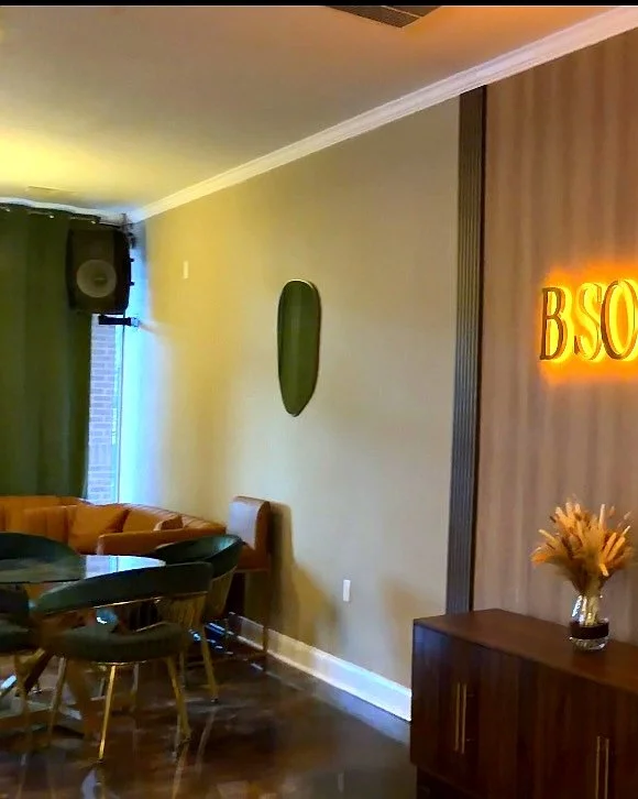 Modern interior room with a beige sofa, a black dining table with matching chairs, a green curtain, and decorative elements including a wall art piece, a lit-up sign reading 'BSO,' and a vase with dried flowers on a wooden cabinet.