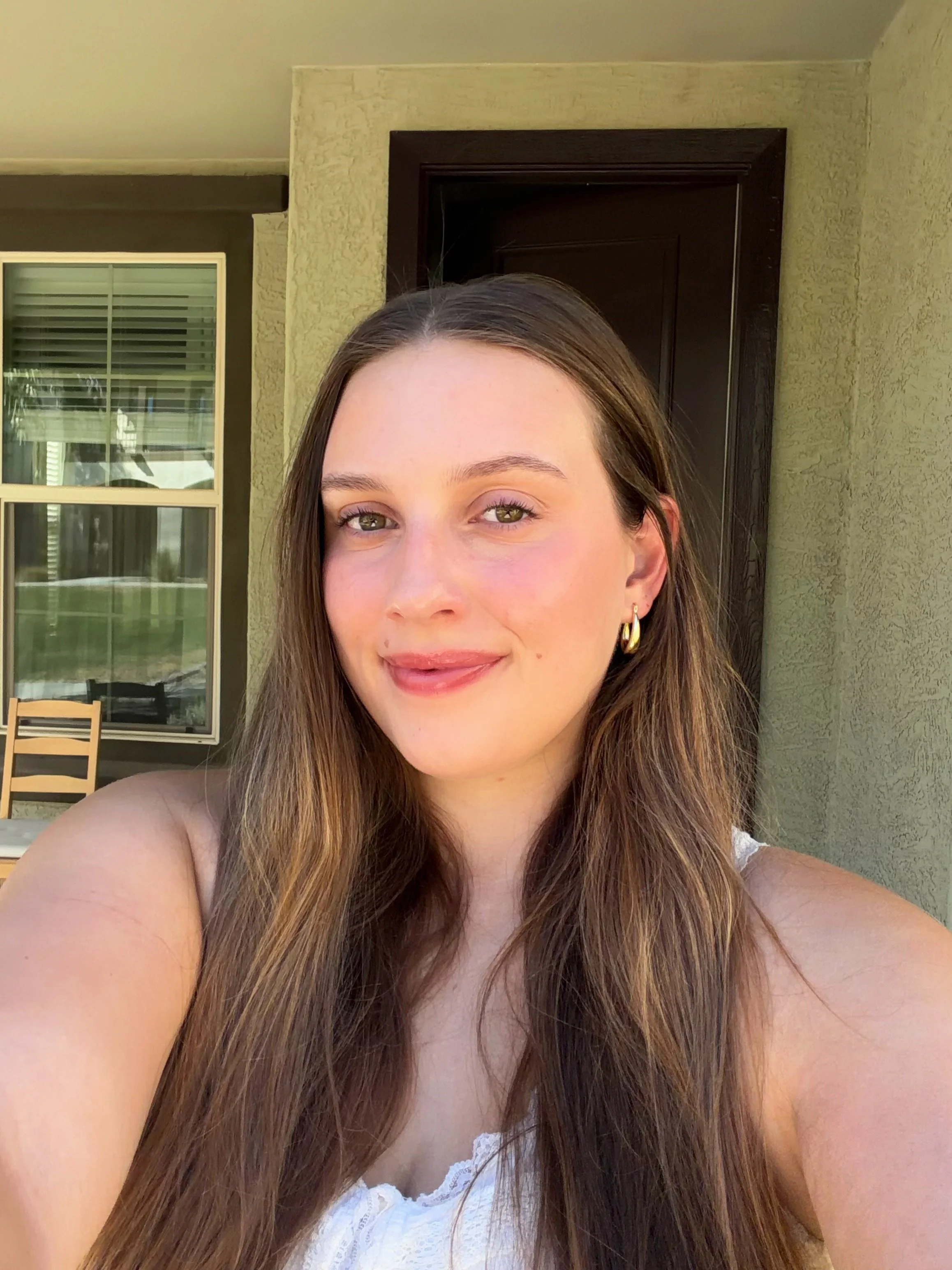 A young woman with long brown hair, green eyes, light makeup, and gold hoop earrings, smiling outdoors on a sunny day in front of a house with large windows and beige walls.
