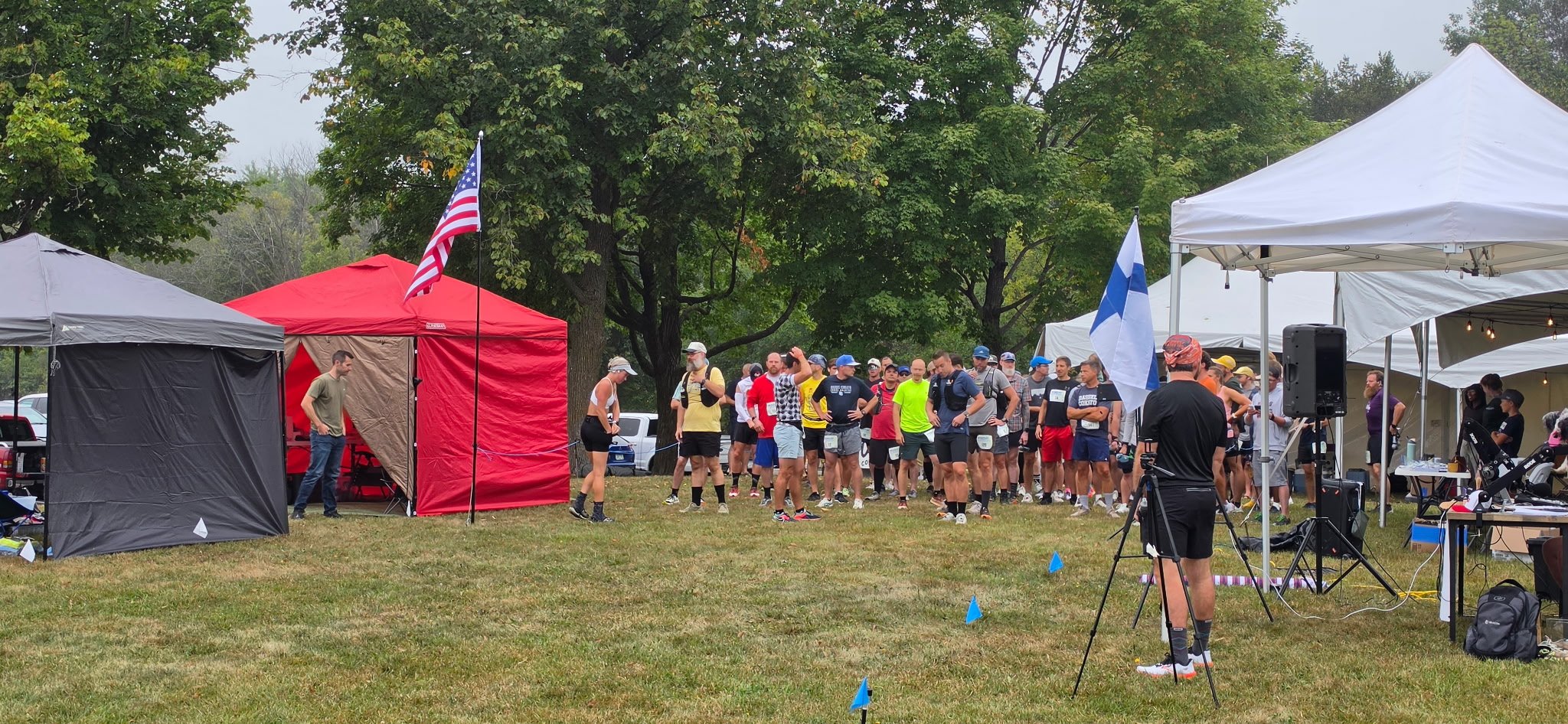 A group of runners gather at the starting line of a race event outdoors, with tents, flags, and green trees in the background.