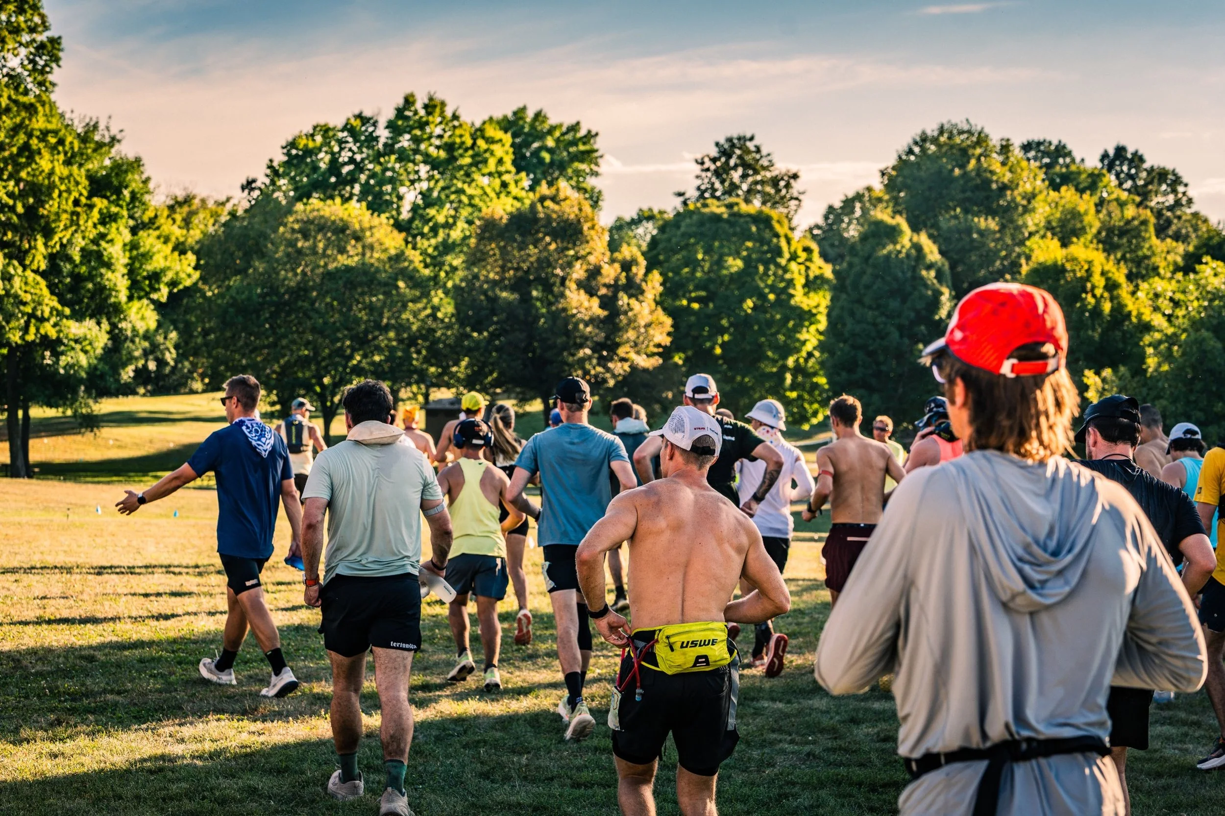 Group of people participating in an outdoor running event in a park during late afternoon, with green trees and a partly cloudy sky in the background.
