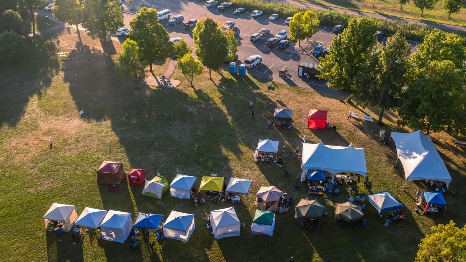 An aerial view of a park with numerous tents set up for an outdoor event, with a parking lot filled with cars and trucks in the background, surrounded by trees and grassy areas.