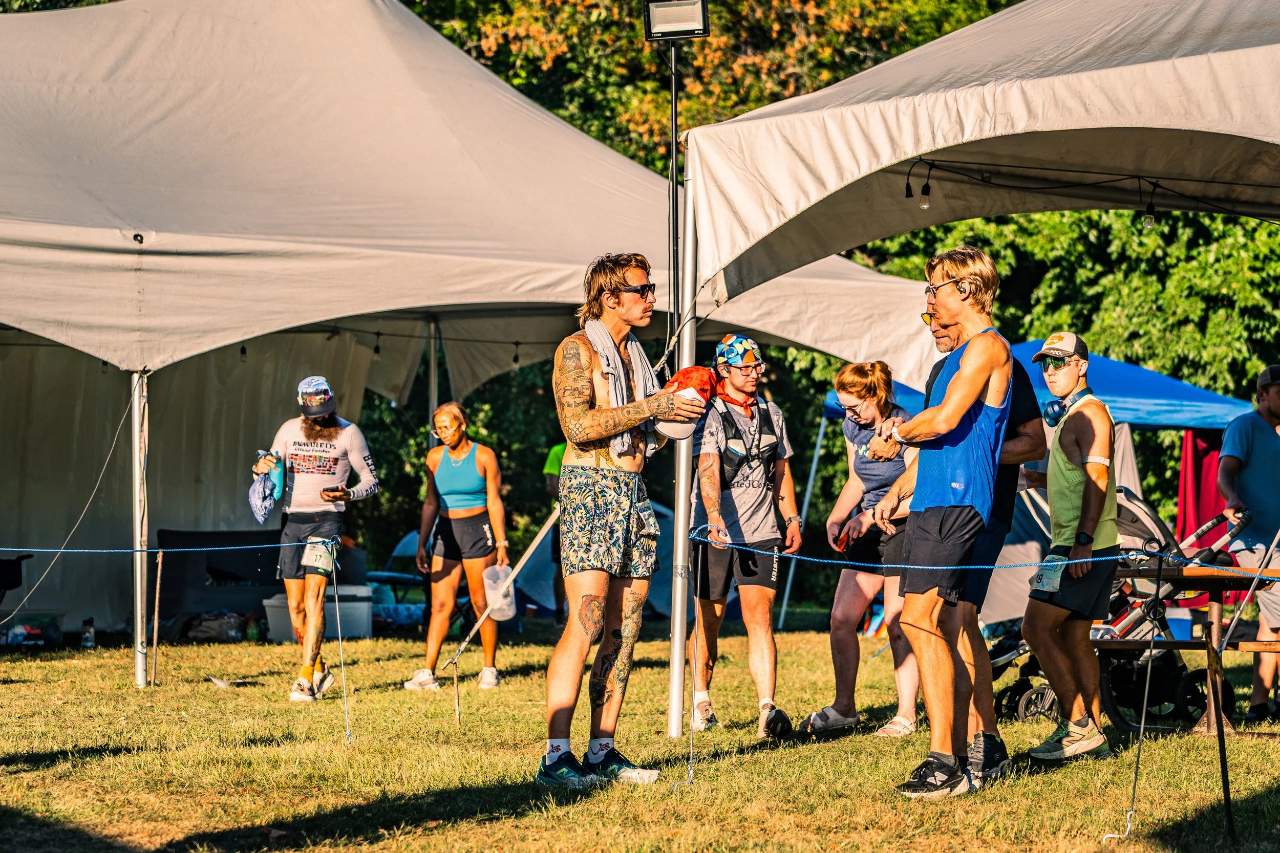 Group of people at an outdoor event, some wearing athletic clothing and gear, under white tents on a grassy area with trees in the background.