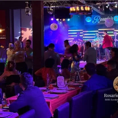 People dining at tables in a restaurant or bar with a live music stage set up in the background, decorated with colorful lights and a blue sign reading 'Rosario's'.