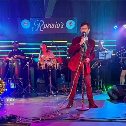 A musical performance on stage at Rosario's in Toronto, featuring a male singer in a red jacket, with band members playing percussion and guitar, illuminated by colorful stage lights.