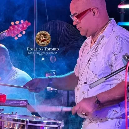 A DJ wearing glasses and a floral shirt is mixing music at a club or party, with a colorful, neon-lit background.