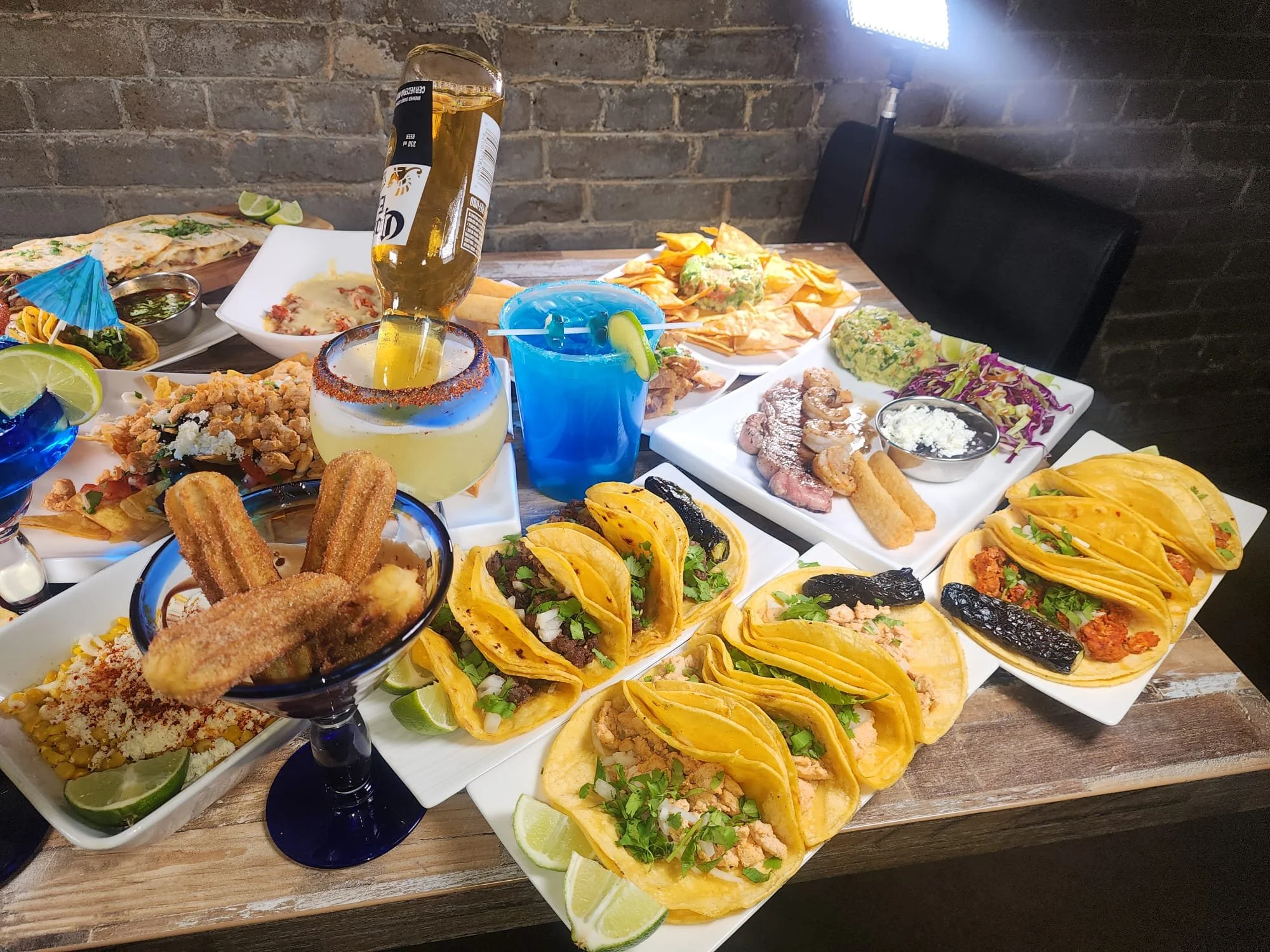 A table filled with Mexican cuisine including tacos, nachos, ceviche, chile rellenos, and various garnishes, with a bottle of beer, a margarita, and a blue cocktail.
