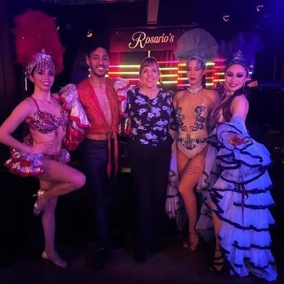 Five people standing on stage at Rosario's, four women in elaborate showgirl costumes and one woman in casual clothing, with a purple and pink backdrop.