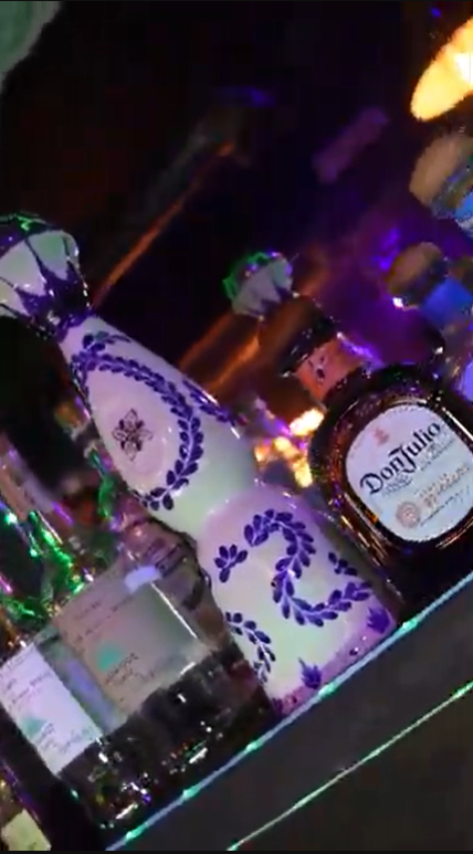 A collection of bottles including Don Julio tequila, a ceramic bottle with purple floral design, and various other alcohol bottles on a bar counter with colorful lighting in the background.