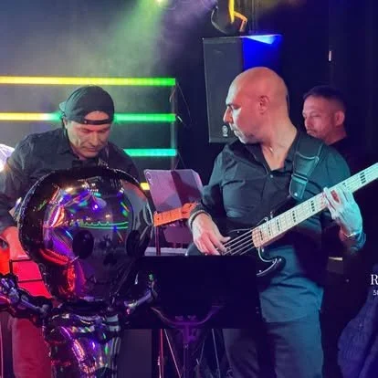 Musicians performing live on stage with colorful lighting, including a man playing an electric guitar, another man wearing headphones, and a robot-like figure in the foreground.