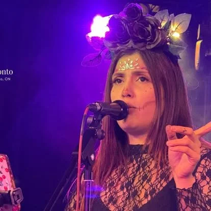 Woman singing into a microphone, wearing a floral crown and lace top, with face paint and stage lighting in the background.