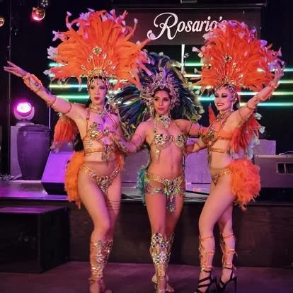 Three women in colorful, elaborate feathered costumes perform on stage with their arms raised in a dance pose.