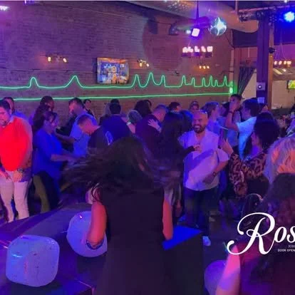 People dancing and socializing in a nightclub with purple and blue lighting, a neon heartbeat line on the wall, and a DJ booth in the background.