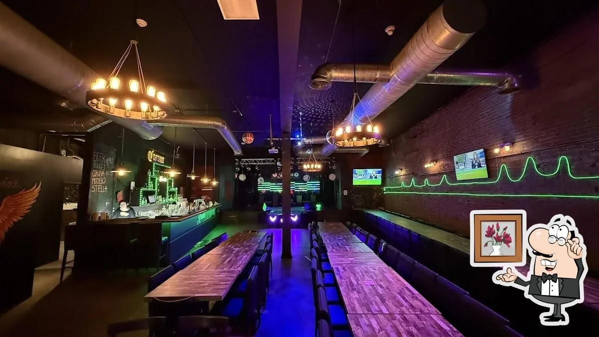 Empty restaurant or bar with long wooden tables, neon lights, TV screens on the wall, and a bar counter with staff behind it.
