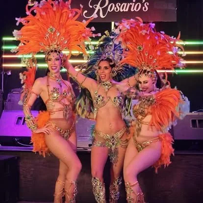 Three women in vibrant, jeweled costumes with large orange feathered headdresses, performing on stage with colorful lights.