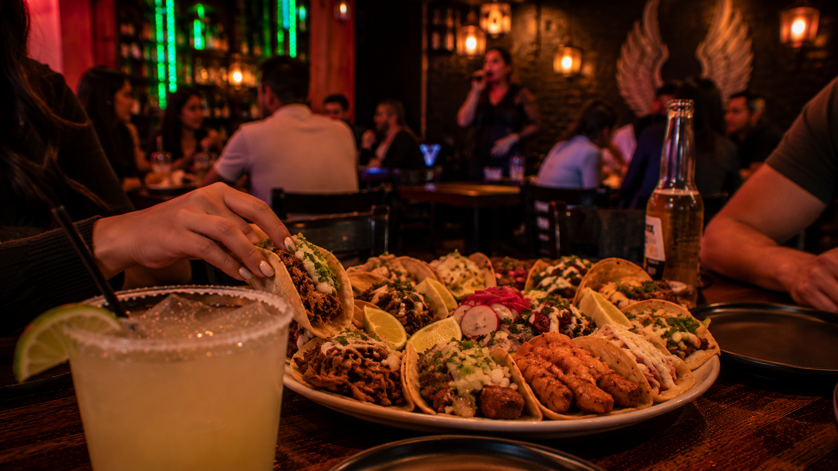 A plate full of assorted tacos with lime wedges, in a lively restaurant. A woman is holding a taco, and there is a margarita with a lemon wedge in the foreground. In the background, people are dining and a performer is singing on stage.