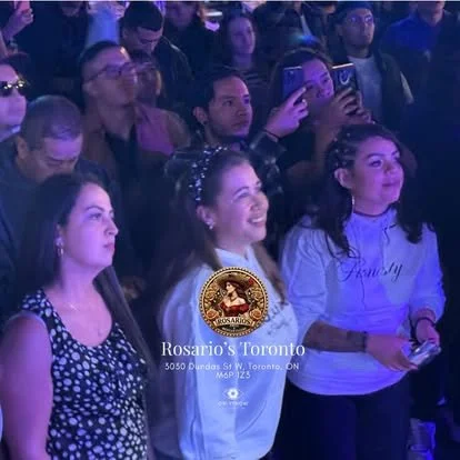 Audience at a concert or event, mostly women, some taking photos or videos with smartphones, some smiling or watching attentively, with a logo and location info at the bottom.