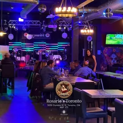 Interior of a lively bar or nightclub with a band stage in the background, colorful lighting, patrons sitting at tables, and a woman standing near the bar.