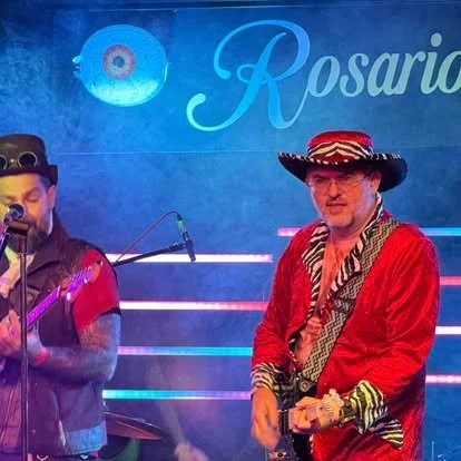 Two musicians performing on stage with a blue sign that says 'Rosario' and a decorative eye symbol in the background. One musician is playing guitar and singing, while the other is wearing a sombrero and traditional Mexican attire.