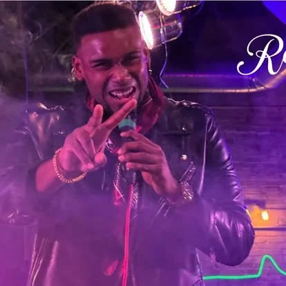 A young man making a peace sign and smiling while performing with a microphone in a dimly lit setting with purple and pink lighting.