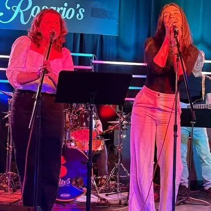 Two women singing on stage with a band in the background, under colorful stage lights.