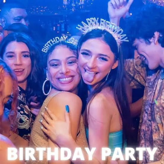 Group of people celebrating a birthday party at a lively event with festive accessories and colorful lighting.
