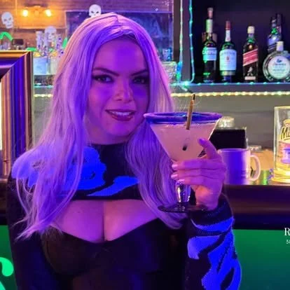 A woman with long blonde hair smiling and holding a cocktail in a bar with colorful bottles and neon lights in the background.