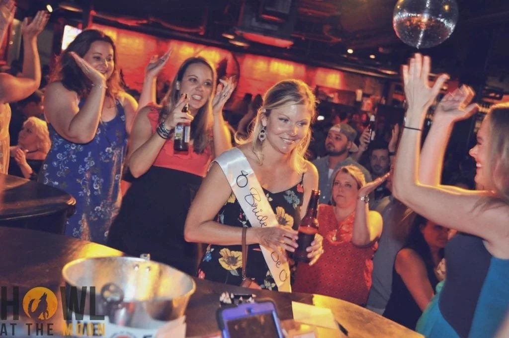 A group of women celebrating in a bar, with one woman wearing a sash and holding a beer, surrounded by friends dancing and smiling.