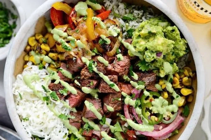 Bowl of Mexican salad with grilled beef chunks, rice, corn, guacamole, shredded cheese, and sliced red onions with drizzle of green sauce.