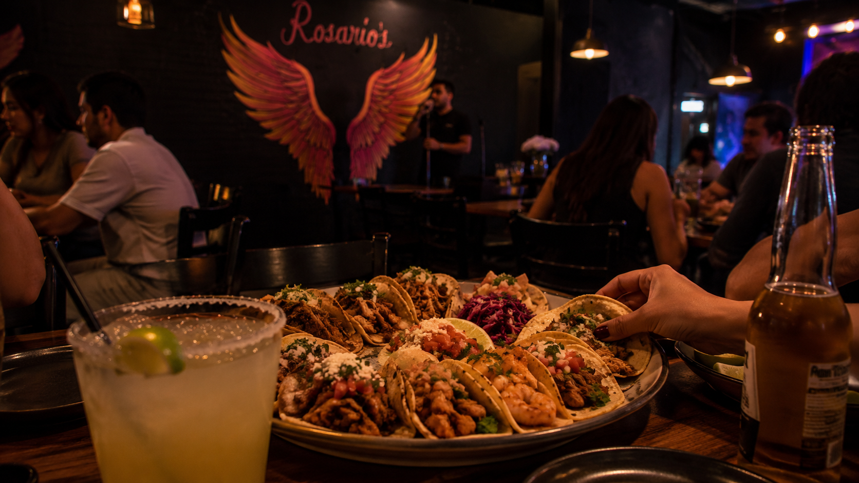 Plate of tacos on a table at a dimly lit restaurant with people dining and a person on stage speaking in the background.