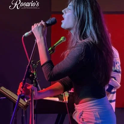 A woman singing into a microphone at a performance venue with a sign that says 'Rosario's' in the background.