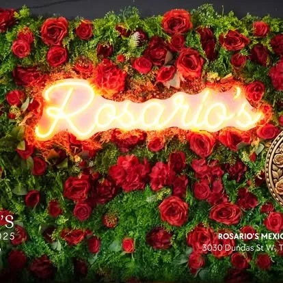A floral arrangement with red roses and green foliage, featuring a neon sign that reads 'Rosario's' in cursive.