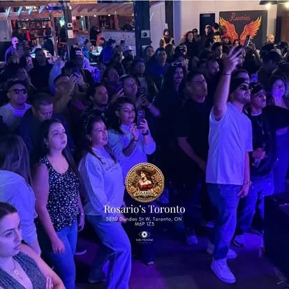 A crowd of people at a concert or event, some dancing and enjoying the music, with purple lighting and a banner that says 'Rosario's Toronto' in the background.