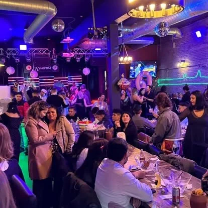 People socializing and dining at a lively indoor venue with colorful lighting, disco balls, and a stage or performance area.