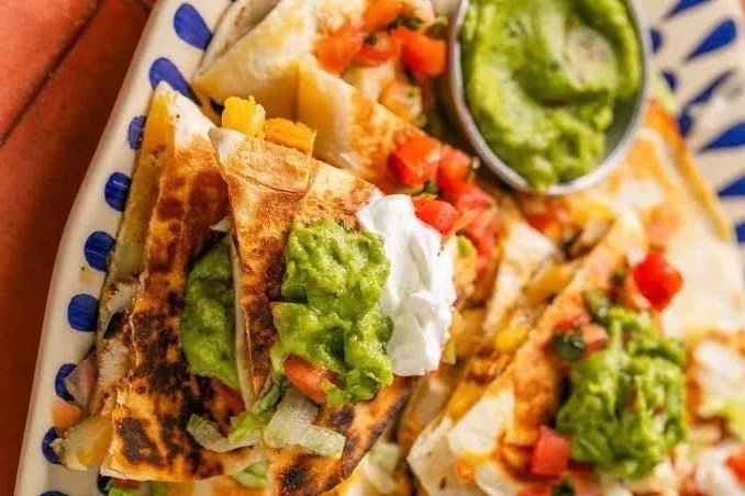 Close-up of two beef tacos with shredded lettuce, diced tomatoes, sour cream, and guacamole, served with a side of guacamole on a decorative plate.