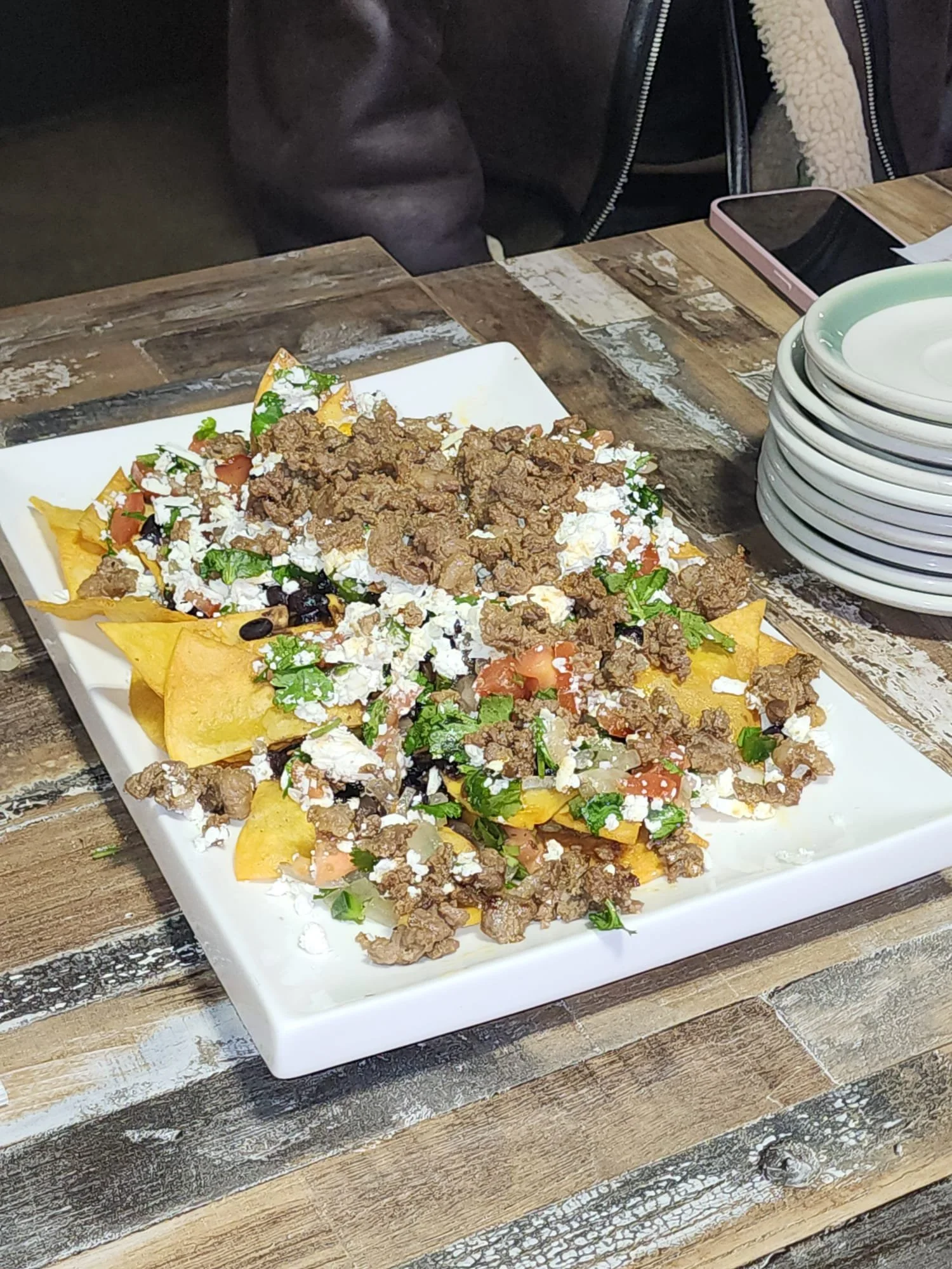 A plate of nachos with beef, cheese, tomatoes, black beans, cilantro, and sour cream, placed on a rustic wooden table.