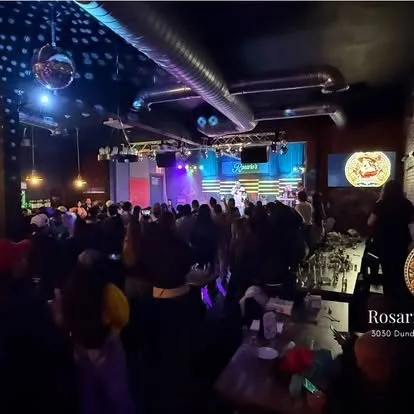 Crowd gathered at a nightclub for a live performance on stage with colorful lights and screens, with a bar in the foreground.