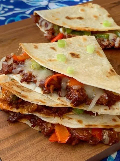 Stacked beef and vegetable tacos on a wooden surface, filled with ground beef, cheese, lettuce, tomatoes, and green onions, with flour tortillas.