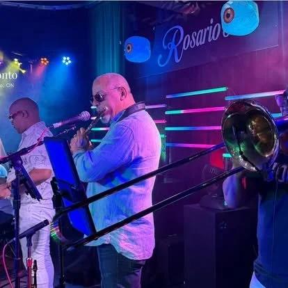 Musicians performing with a saxophone and trombone on stage at a venue called Rosario, with colorful lighting and decorative elements.