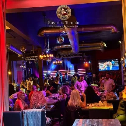Inside Rosario's Toronto restaurant with people dining, a live music stage, and a television showing a boxing match.