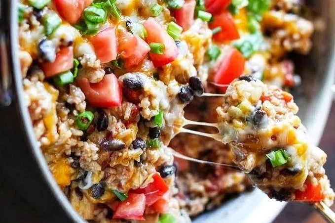 Close-up of a skillet of cooked taco casserole with tomatoes, black beans, corn, cheese, and green onions, with a serving spoon lifting a portion.
