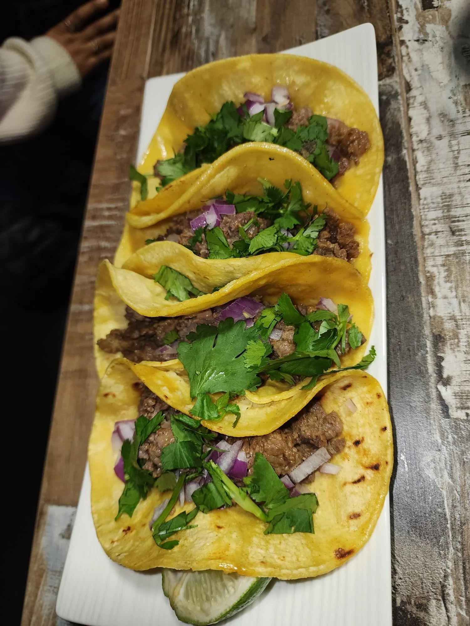 Three tacos filled with seasoned ground beef, topped with chopped red onions and chopped cilantro, served on a white rectangular plate with a lime wedge on the side.