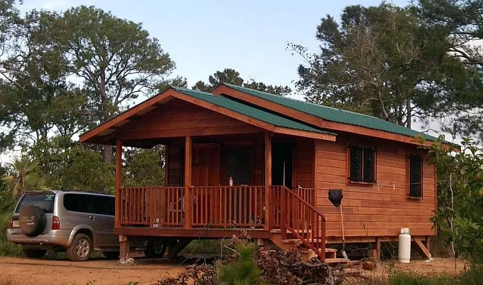 Small wooden house on stilts with a green roof, front porch, and steps leading up to it, surrounded by trees and dirt ground, with a silver van parked nearby.