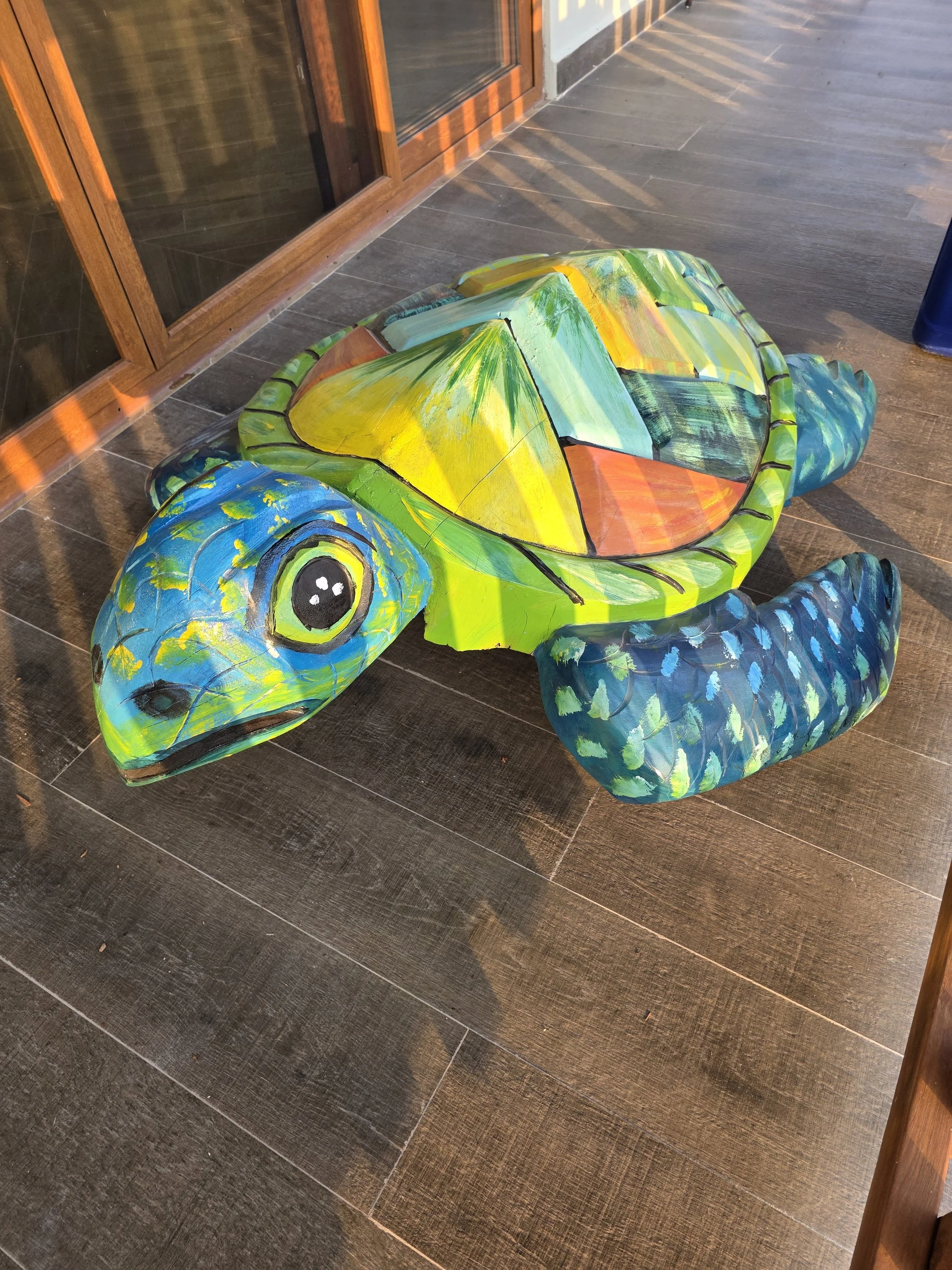 Painted wooden turtle sculpture with colorful geometric patterns on its shell, placed on a floor near glass doors.